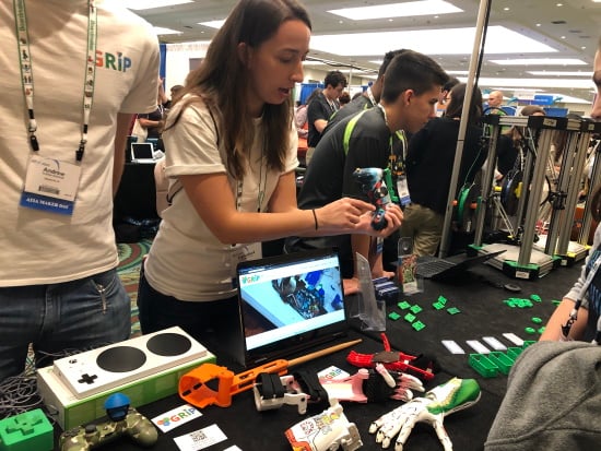College students at a table of 3D printed devices including prosthetic hands and switches. Some are hand painted with themes. 3D printers are visible on the far right.