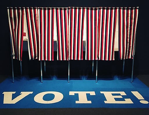 Four voting booths with red, white and blue stripped curtains on a carpet with the word VOTE! in capitals