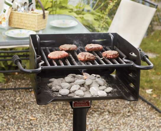 A grill on a post with a grate and side handles. Burgers are cooking above burning briquettes.