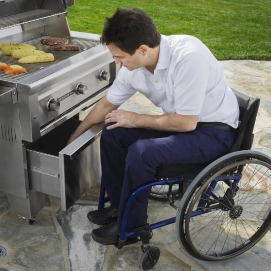 A man in a manual wheelchair reaches into a deep drawer under a gas grill.