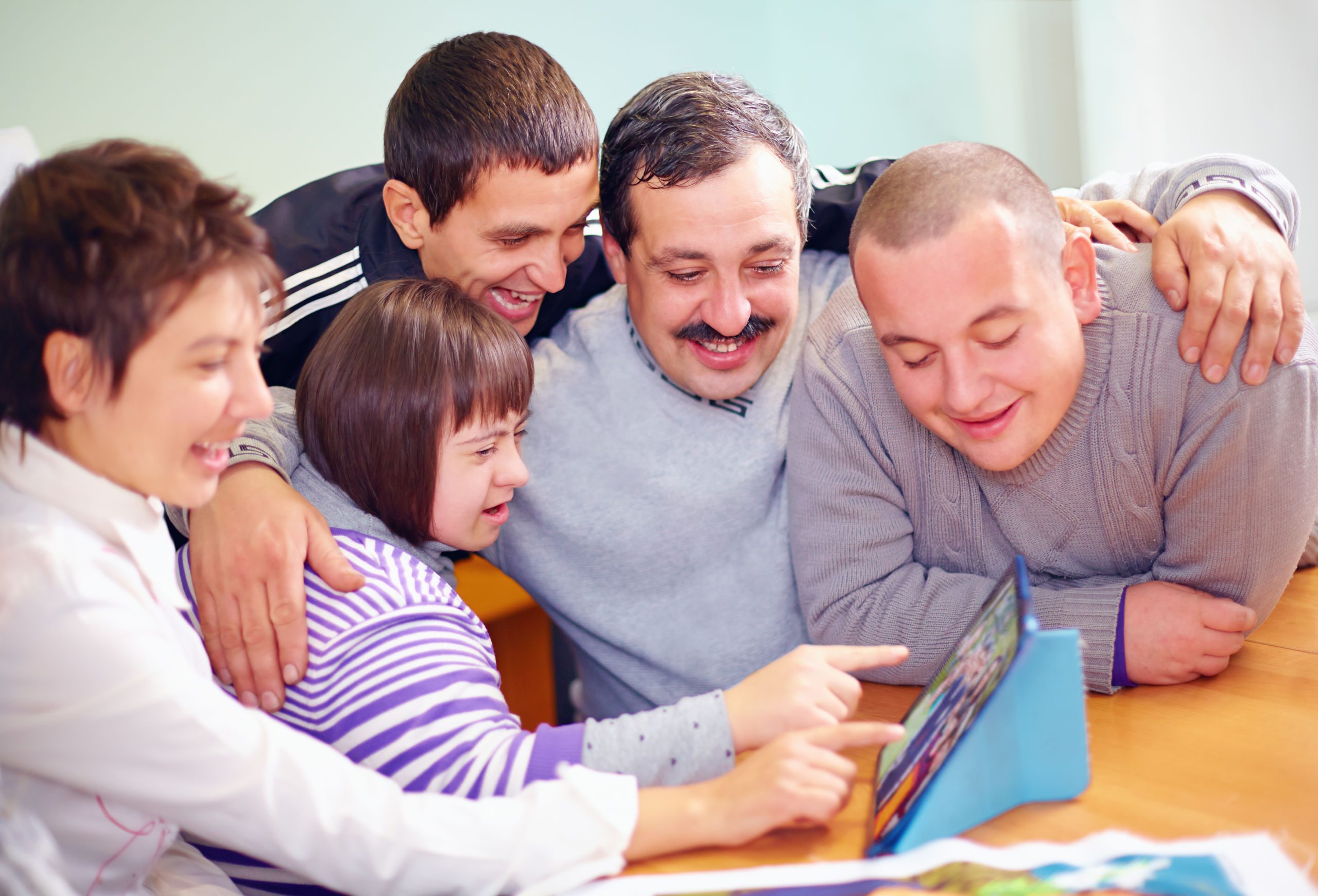 A family of five smiling and clustered over a tablet computer. The daughter has a developmental disability and is pointing at the screen with her mother.