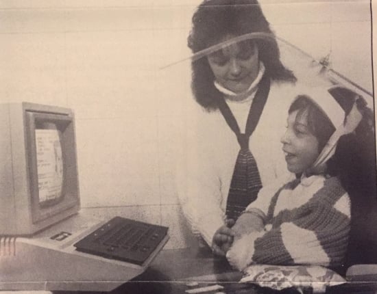 A little girls wearing a head pointer smiles at an early desktop computer. A woman stands beside her.