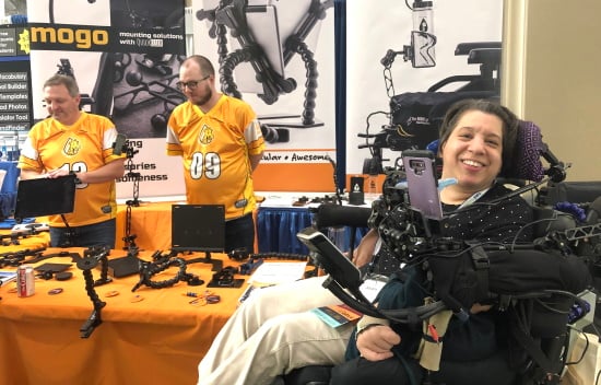 A woman smiles from her power chair in front of a vendor booth selling wheelchair mounting equipment.