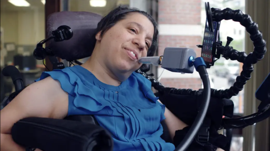 A smiling woman seated in a power wheelchair mounted with a smartphone and mouth-controlled joystick controller.