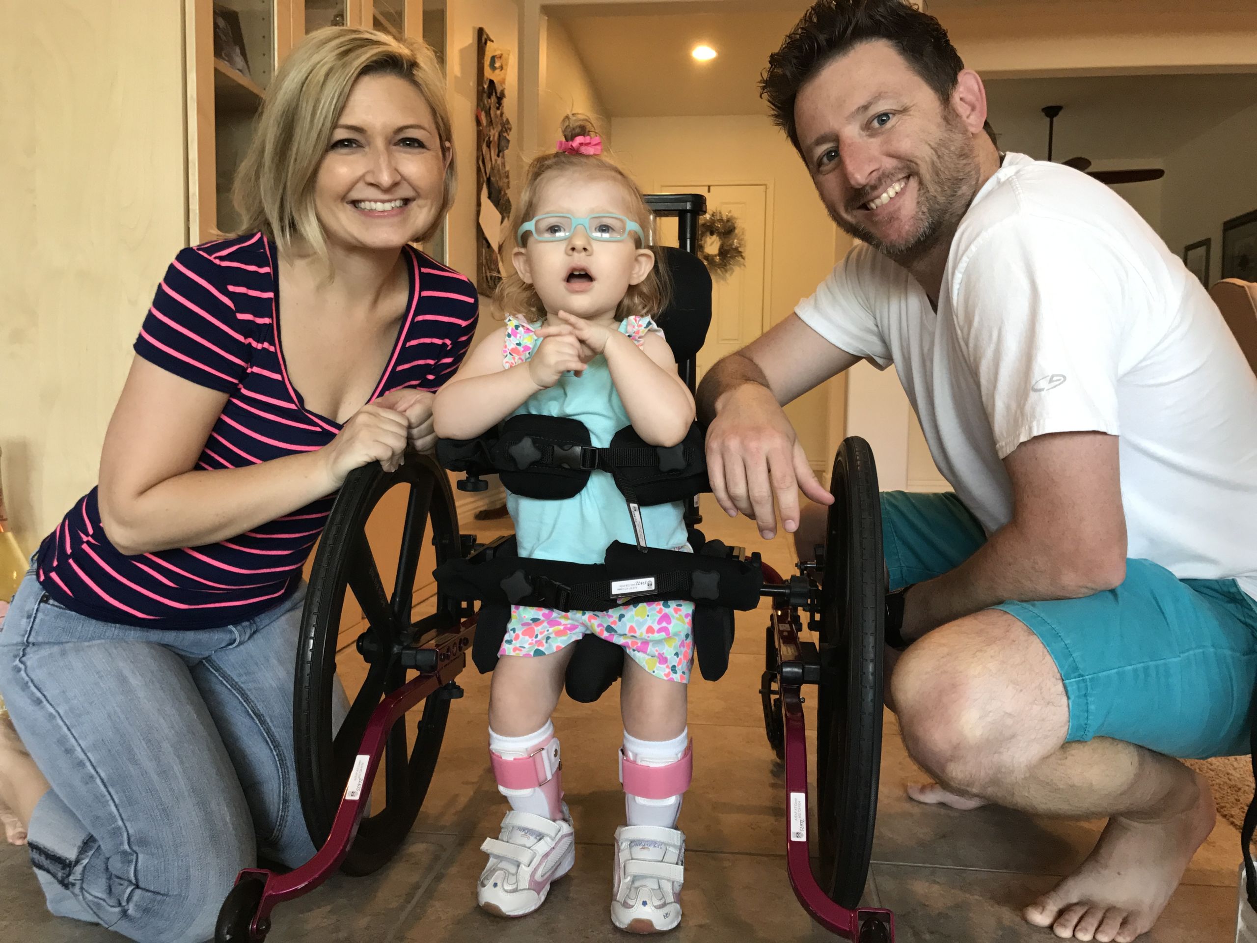 A little girl standing supported by a wheeled gait-trainer device for walking and flanked by her smiling parents.