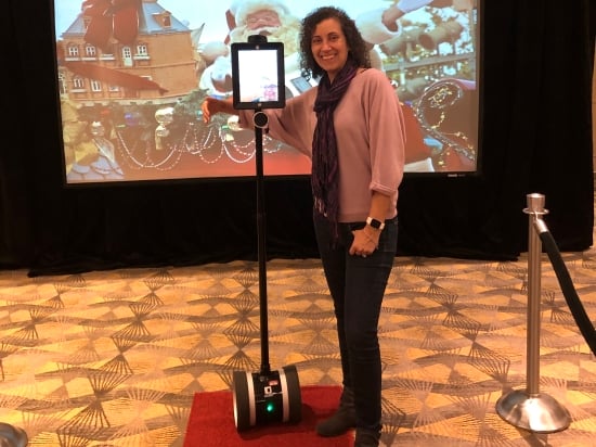 A woman in a convention hall stands smiling next to a robot that resembles a tablet computer mounted on Segway wheels.