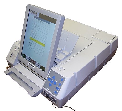 An electronic box that resembles a printer, but has a large touch-screen tablet facing the user. Beneath is a feed tray for inserting a paper ballot. On the right side are buttons for tactile navigation and beneath are jacks for headphones and other devices.