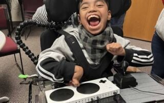 A smiling child sitting in a wheelchair and using a switch adapted game controller and joystick