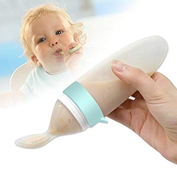A soft bottle filled with pureed food that dispenses to a silicone spoon held by a hand. A baby is in the background.