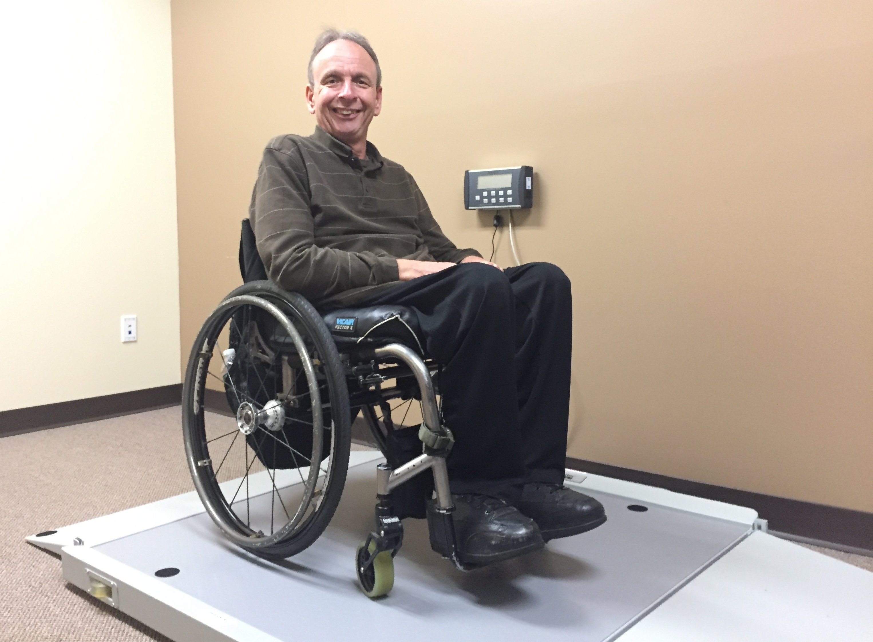 Smiling man in manual wheelchair on a roll-on scale in a small room.