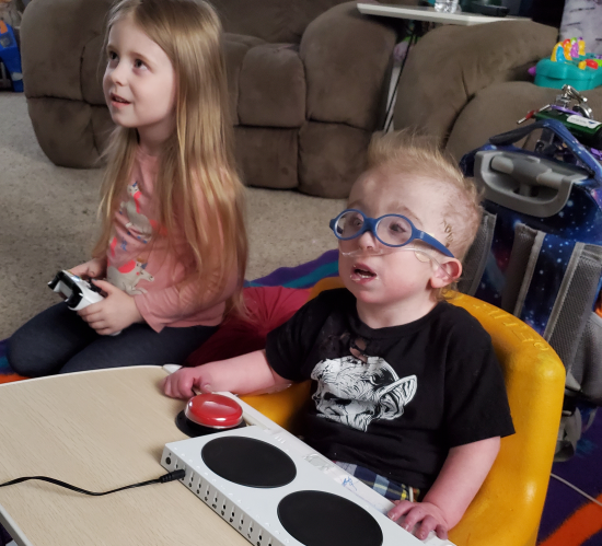 A little boy wearing oxygen tubing sits on the floor using an adapted Xbox controller and switch. A little girl kneels next to him holding a conventional controller.