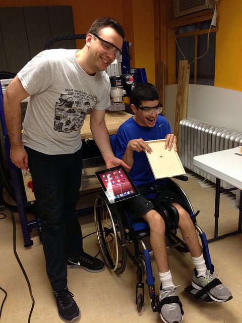 Two men in a workshop, one seated in a wheelchair smiling with a custom-made iPad case, the other stands smiling with the iPad.