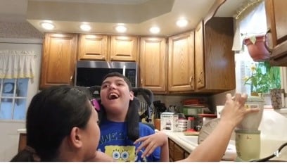A young boy smiling using a switch with his cheek to activate a blender in his kitchen with his mother.