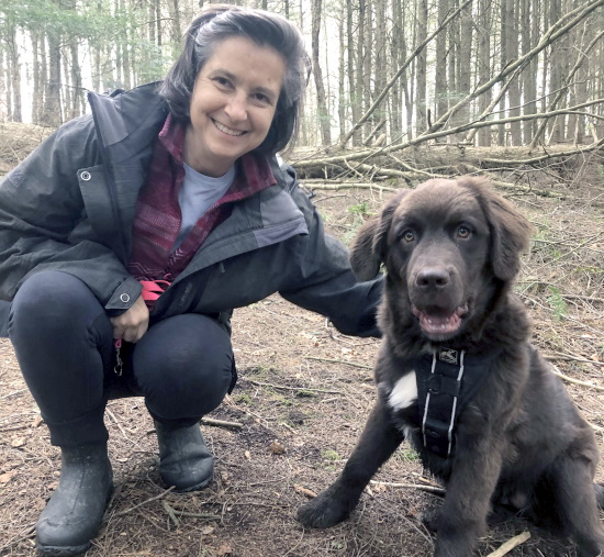 A woman smiles for the camera while crouching next to her puppy in the woods.