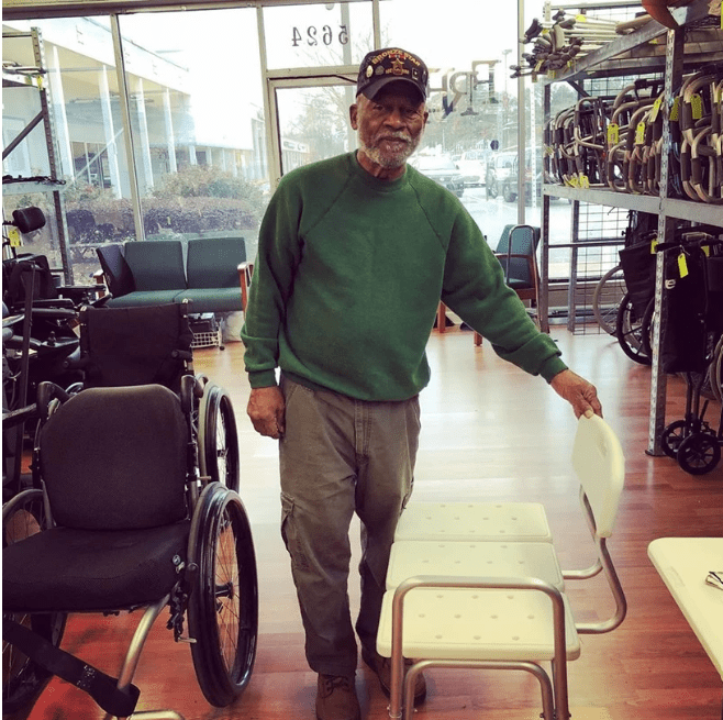A Black man with stands in a show room of durable medical equipment next to wheelchairs, one hand on the back of a transfer bench.