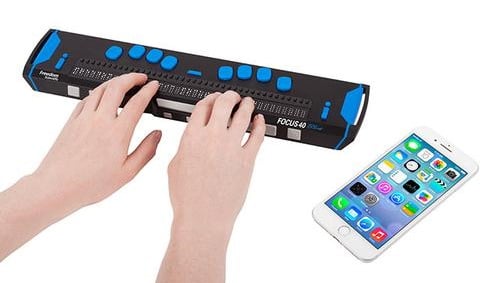 Two hands reading with a Freedom Scientific braille display resting next to an iPhone.