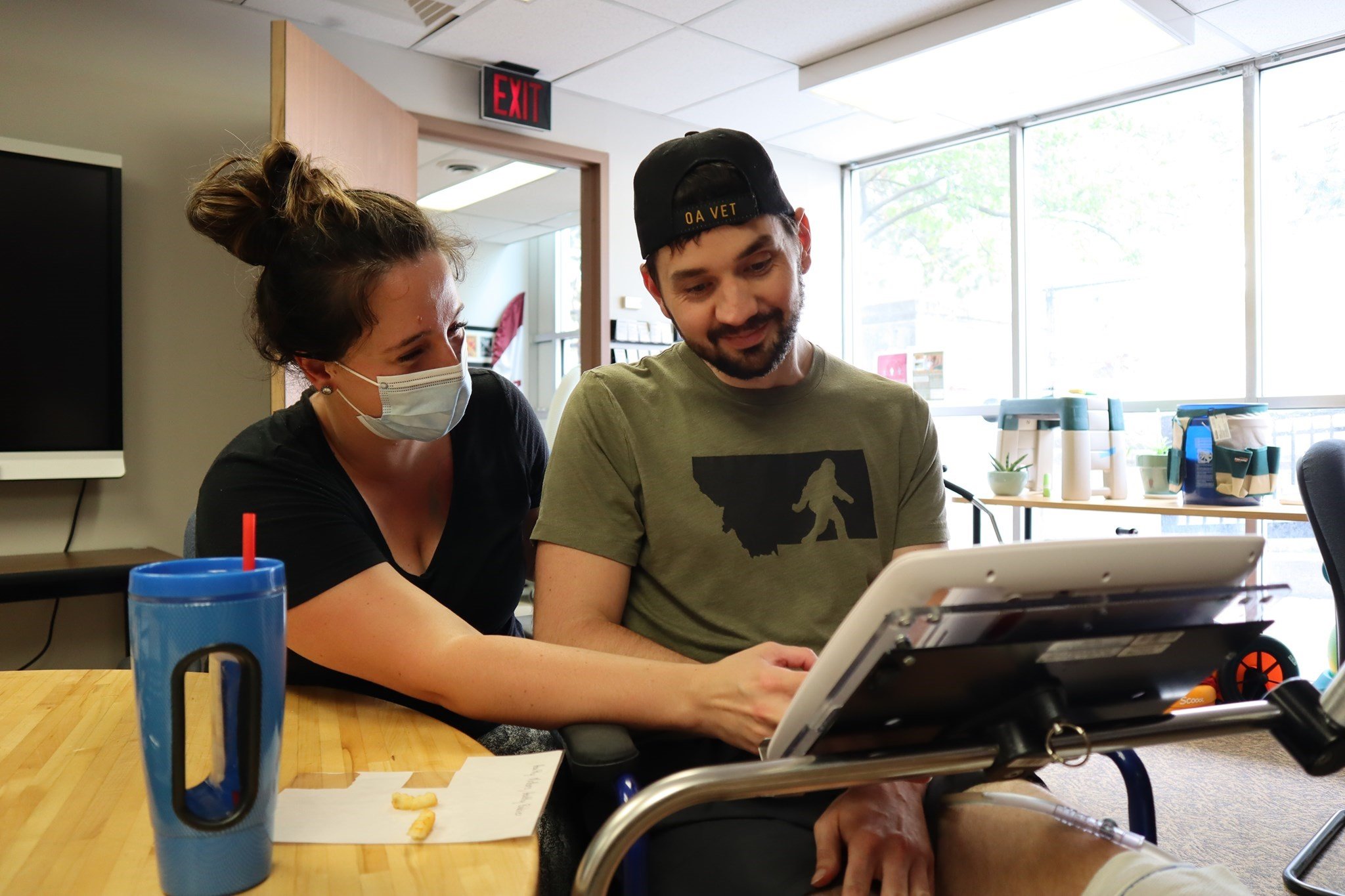 A smiling man in a wheelchair receives help with a mounted tablet computer from a woman smiling behind her mask.