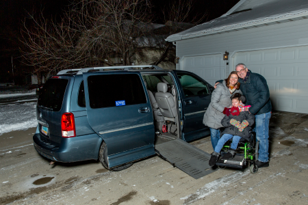 Smiling parents embrace their daughter in a power wheelchair next to an adapted van in front of their garage.