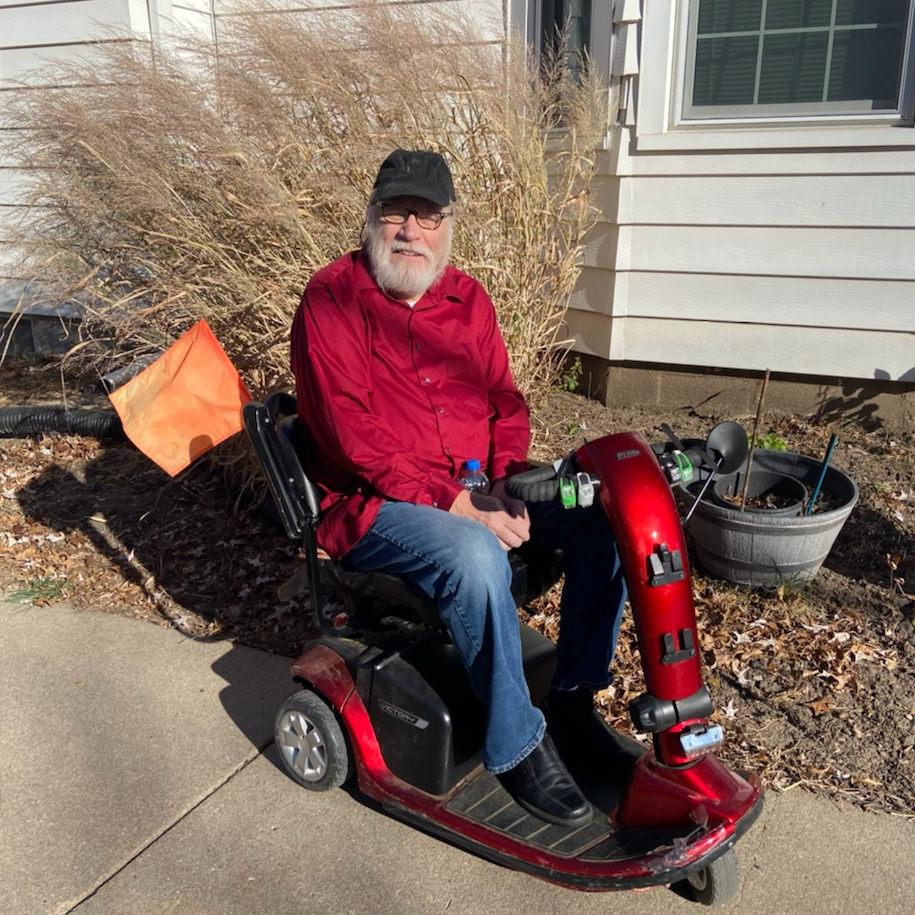 A man with a white beard and mustache smiles seated on a scooter on the sidewalk in a residential neighborhood.