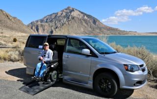 Un hombre sonriente en silla de ruedas eléctrica sale de una furgoneta adaptada en una vista panorámica con montañas y agua.
