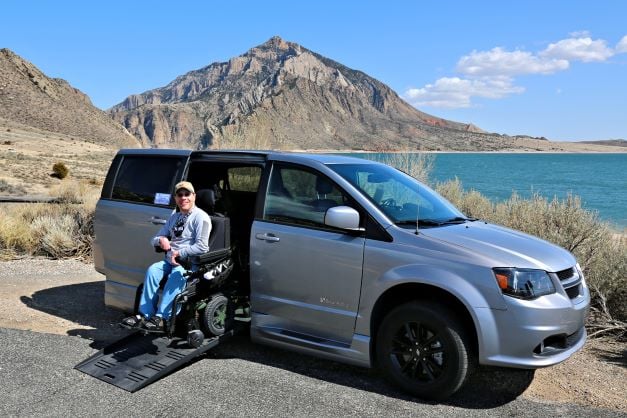 A smiling man in a power wheelchair exiting an adapted van at a scenic vista with mountains and water.