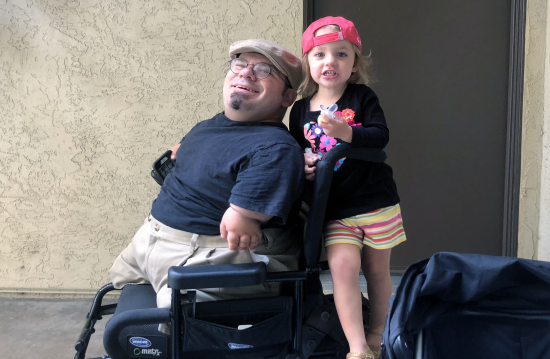 A smiling man seated in a wheelchair with a little girl holding on to the the back.