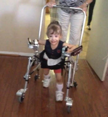 A smiling little girl using a pediatric walker with forearm supports and hip guide. There's an adult guiding from behind.