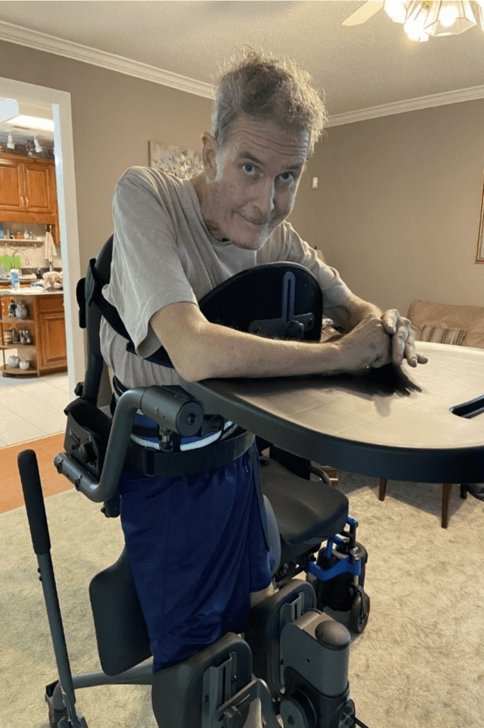 A man at home standing with use of a mechanical stander, arms resting on the device's tray.