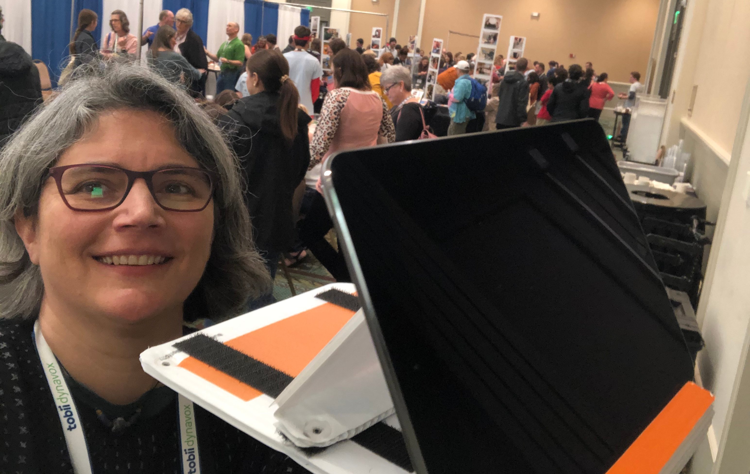 A woman smiles holding her iPad in it's fabricated stand in front of her in the foreground. Behind is a convention all with a crowd of people.
