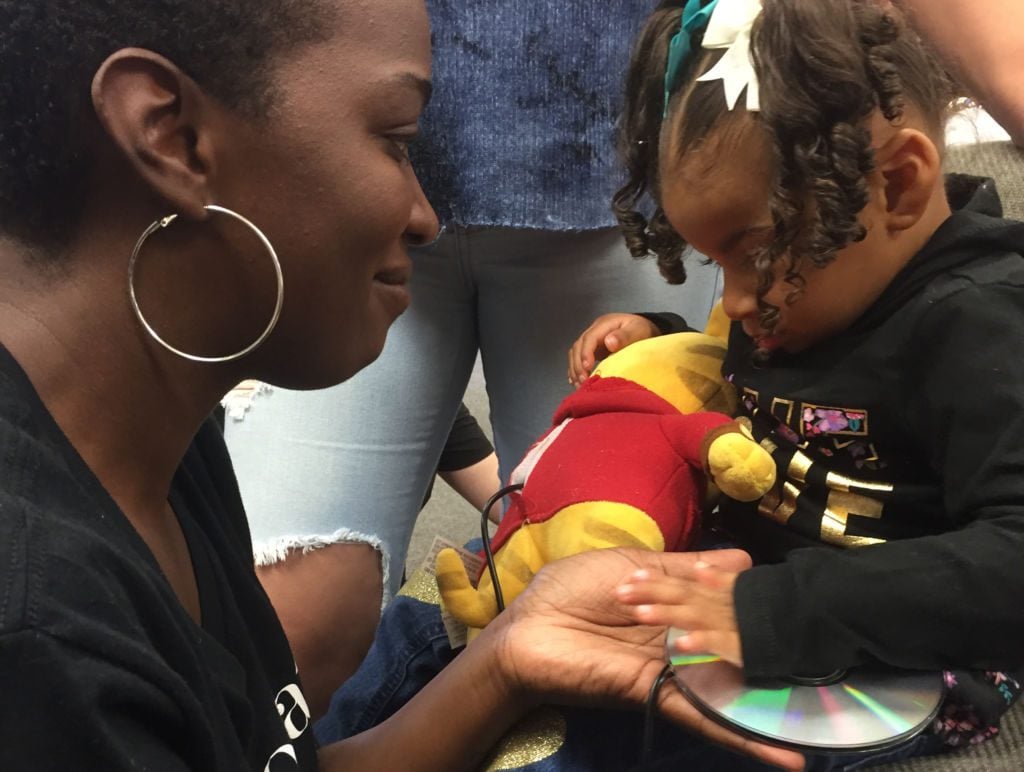 A little girl holding an adapted toy animal presses a CD switch to activate it while her mother looks on smiling.