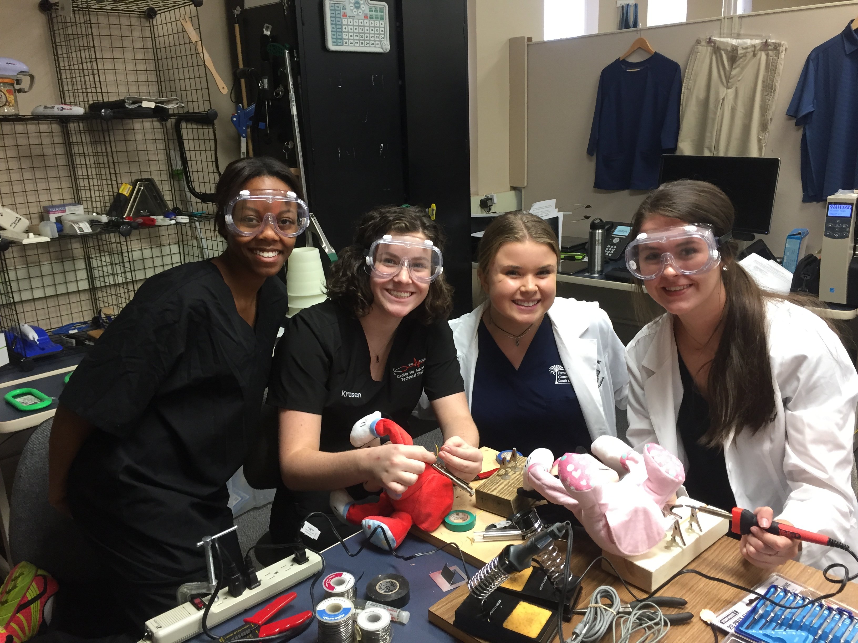 Four young women working at a workbench with soldering irons and plush toys smile for the camera.