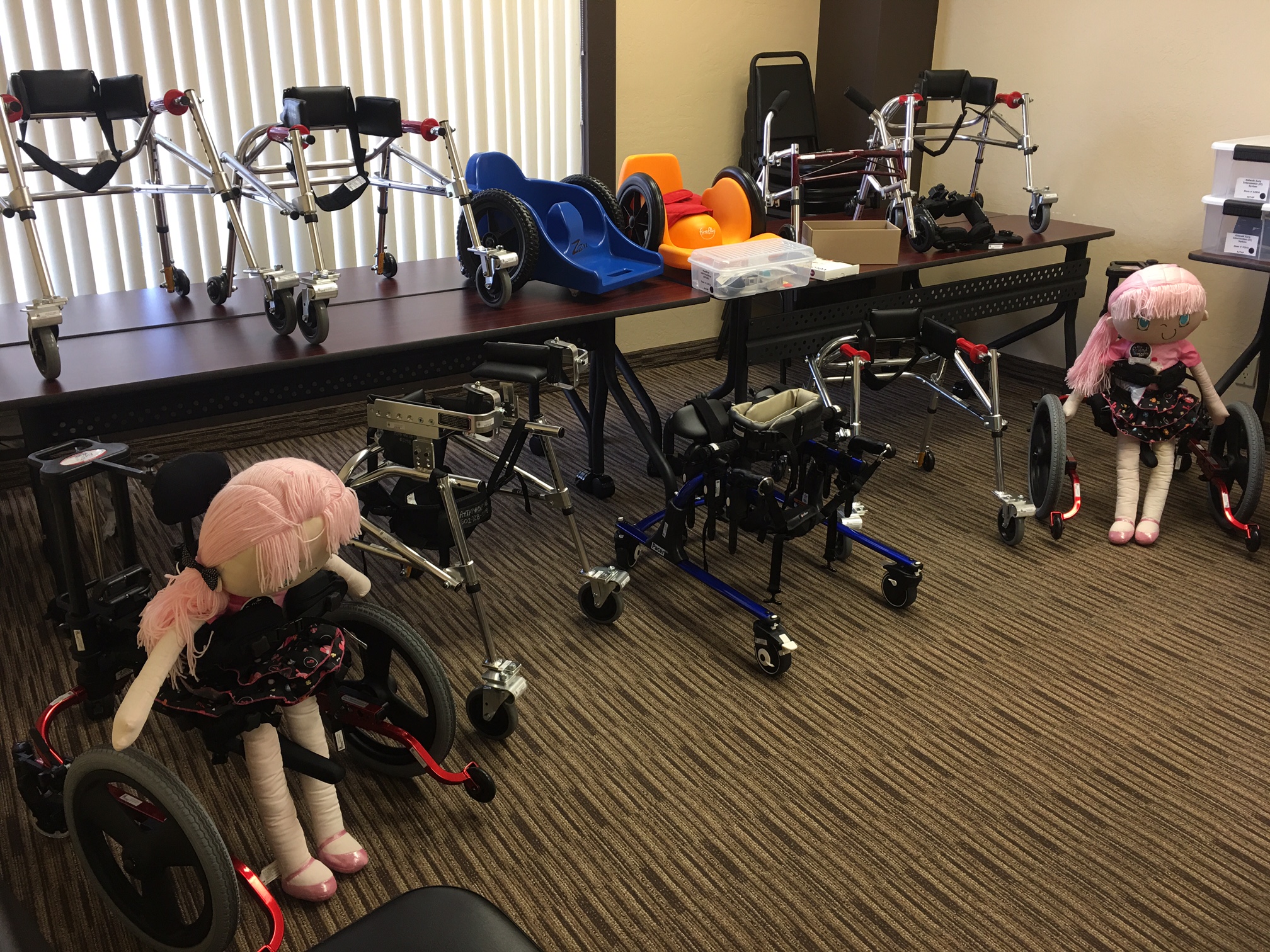 A room full of equipment, mostly walkers and gait trainers. Two support child-size dolls.