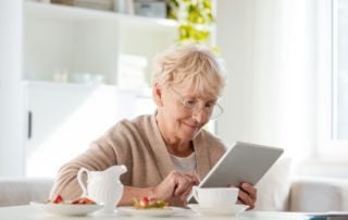 Una mujer blanca de edad avanzada sonríe utilizando una tableta en la mesa de su cocina.