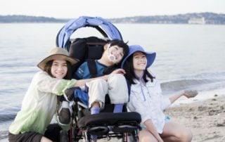 Sisters kneeling and smiling with their brother in an adapted stroller on a beach.