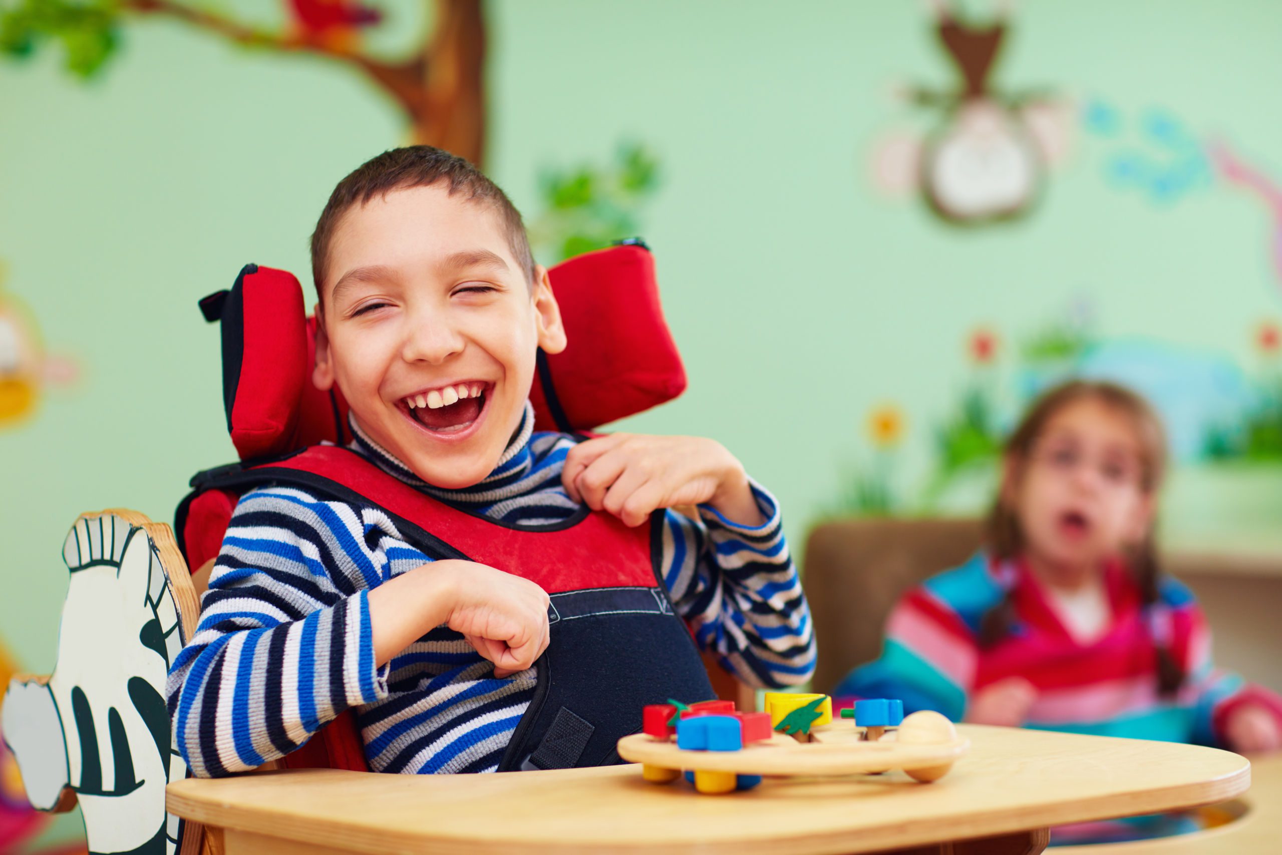 A smiling boy in a classroom seated in an adapted highchair with a puzzle on his tray.