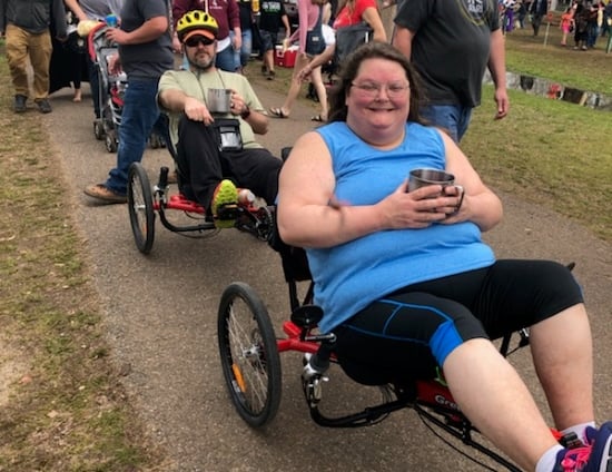A man and a woman seated in recumbent trikes, the woman in front and smiling broadly. They are outdoors on a path with a crowd of people behind them.