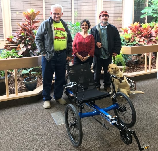 Three adults and a guide dog standing behind a recumbent tricycle.