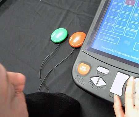 A hand is working the buttons of a tablet-sized voting system with braille beneath buttons and two button switches in contrasting colors beside the tablet.