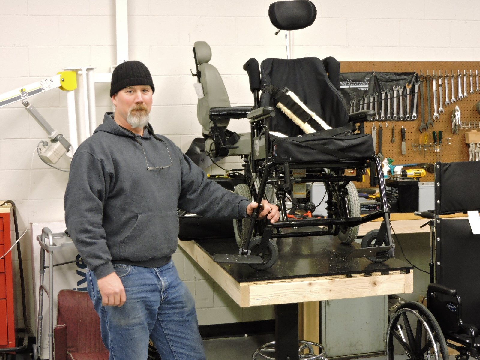 A man wearing jeans and a knit cap stands next to a work bench holding two wheelchairs. Tools are mounted on the wall behind.