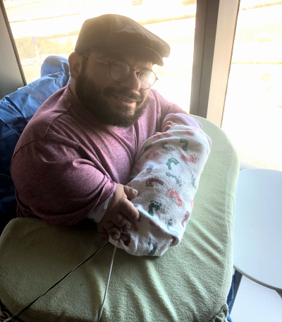A man with short arms holds a baby on a stiff pillow on his lap shaped to wrap around him like a shelf. He is smiling for the camera. The baby is swaddled in a blanket.