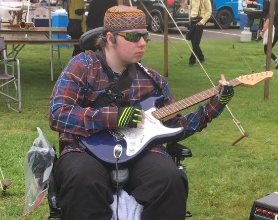 A young man playing an electric guitar seated in a power wheelchair. He is outside and wearing sunglasses and a headset.