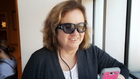 A woman wearing dark glasses with a cord hanging down, holds a smartphone smiling.