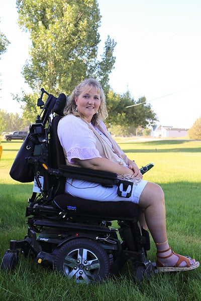 A woman smiling seated in her power wheelchair outside on the grass.