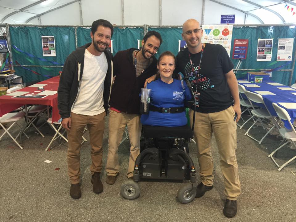A woman in a power chair, who has no arms or legs, smiling, flanked by three men who are standing and smiling. They are in an exhibition tent.