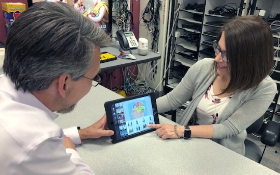 A man and a woman looking at an iPad with an AAC display. In the background are shelves with electronics equipment.