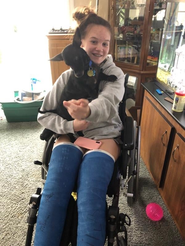 A girl with two broken legs, casts above the knees, seated in a wheelchair smiling with a small dog on her lap.