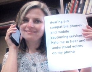 A woman holding a mobile device to her ear holds a sign that reads, Hearing-aid compatible phones and mobile captioning services help me to hear and understand voices on my phone.