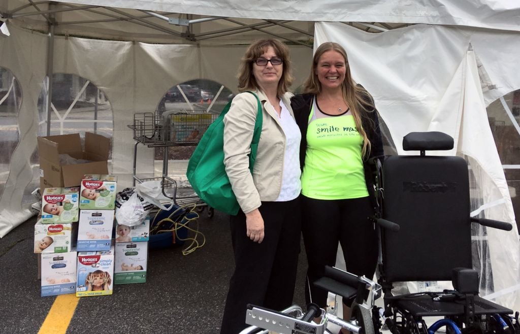 Two women smiling in front of a tent with boxes of adult diapers and next to wheelchairs.