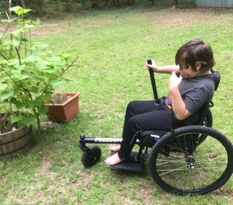 A boy operates the hand levers of an all-terrain wheelchair in a yard with a vegetable garden.