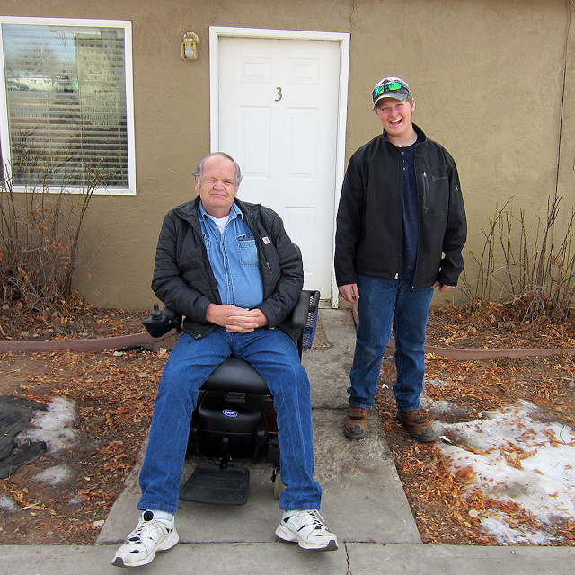A man seated in a power wheelchair smiles next to a smiling teenager standing outside the door to a house.